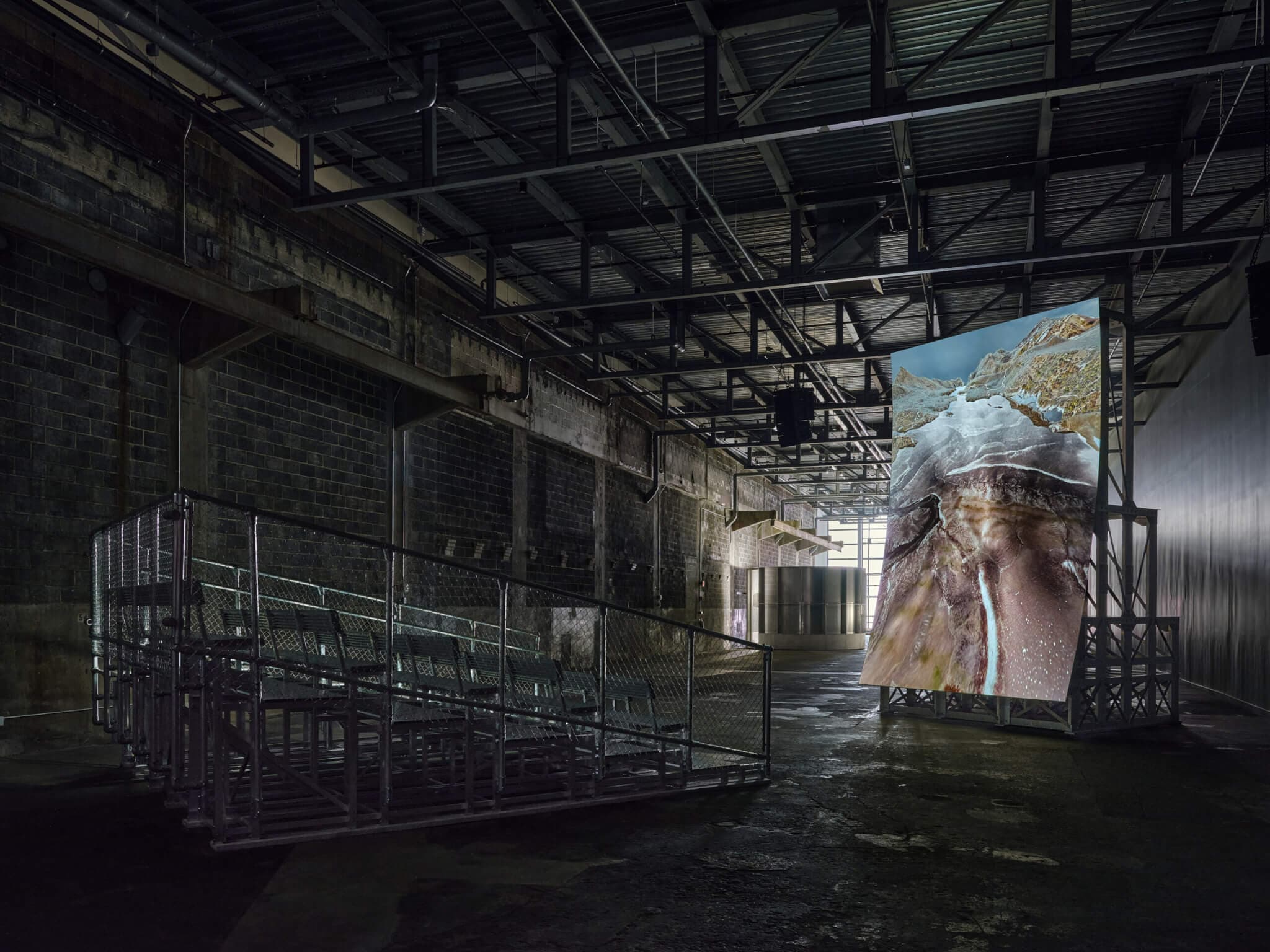 Inside an industrial space, a set of metal bleachers faces a vertical screen displaying an image of rushing water through a cavern.